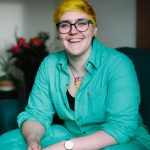 This is a picture of Fiona, a white woman with bright yellow hair. She is smiling and sitting in a relaxed cross legged position, wearing a turquoise suit.