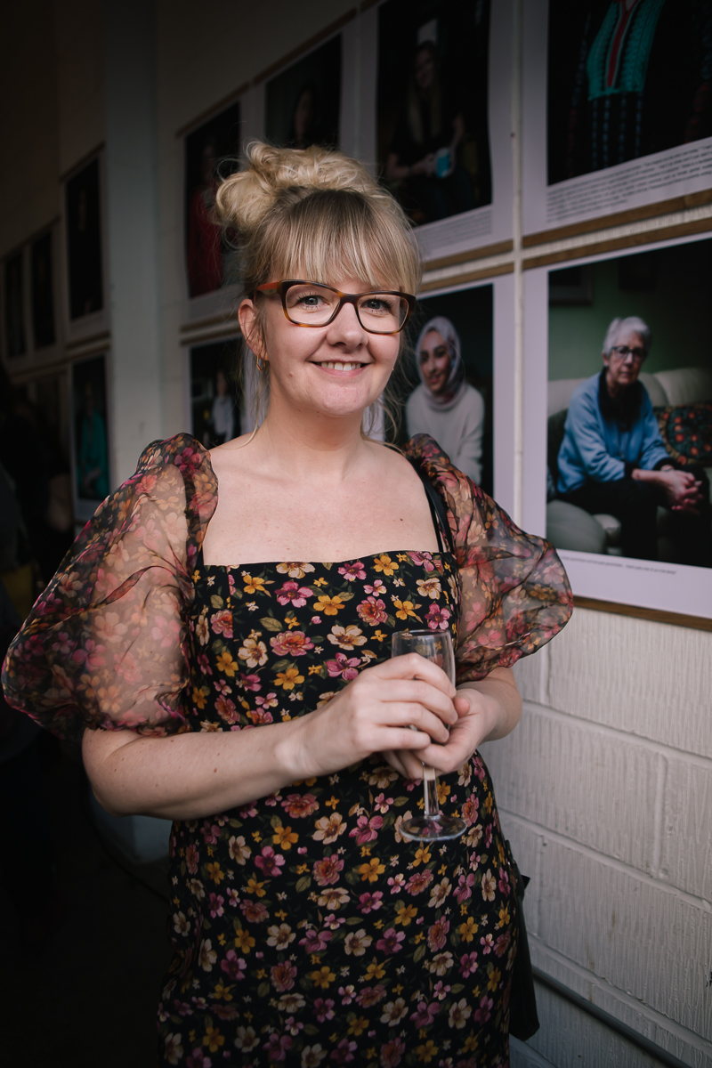A photograph of the artist, Philippa James, a white woman with blonde hair and black glasses, standing in front of a white wall hung with photographic portraits of women. She is wearing a floral dress and holding a glass of sparkling wine. 