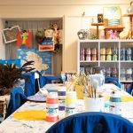 A long table covered in art materials and shelves full of different coloured paints in the AT The Bus art studio.